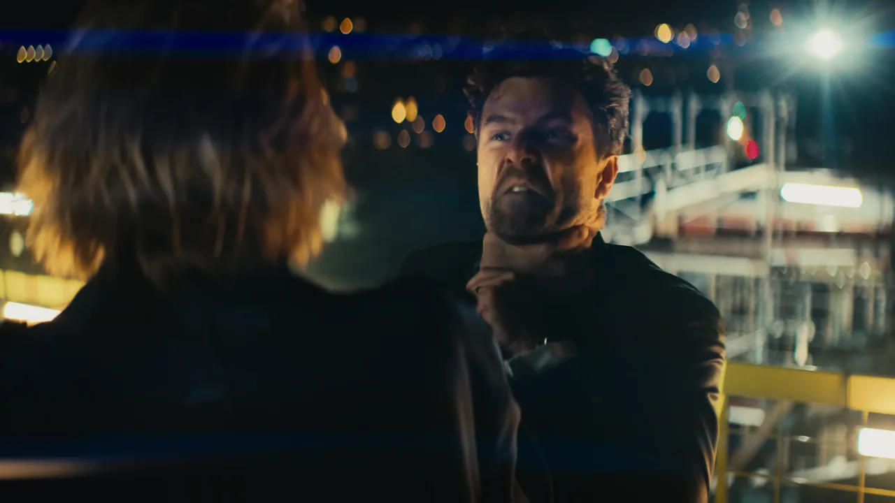 Two men fighting on a rooftop scaffold at night above the city, from the Licence To Sing music video
